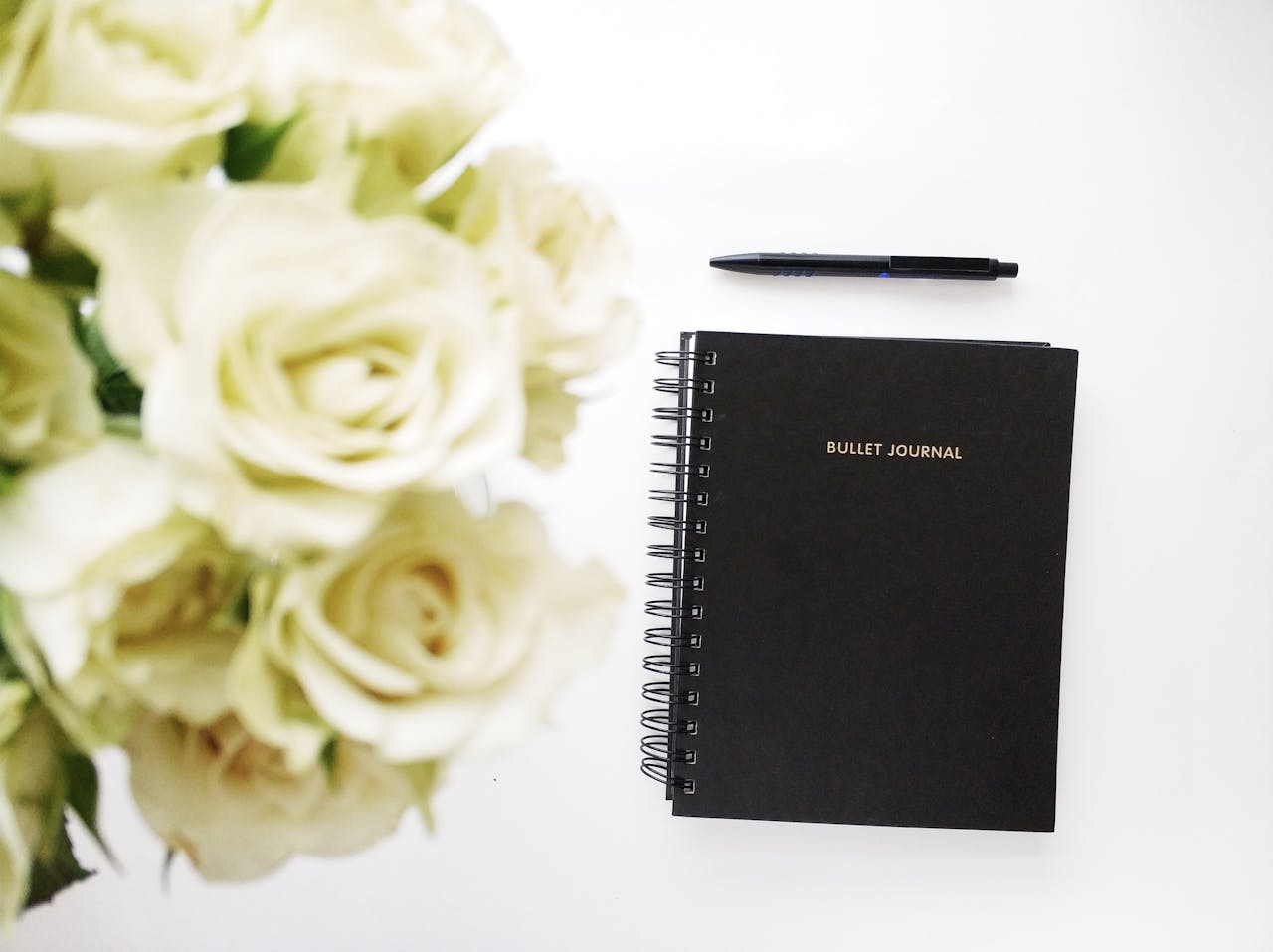 Flat lay of a black bullet journal with a bouquet of white roses, creating a stylish and romantic workspace arrangement.