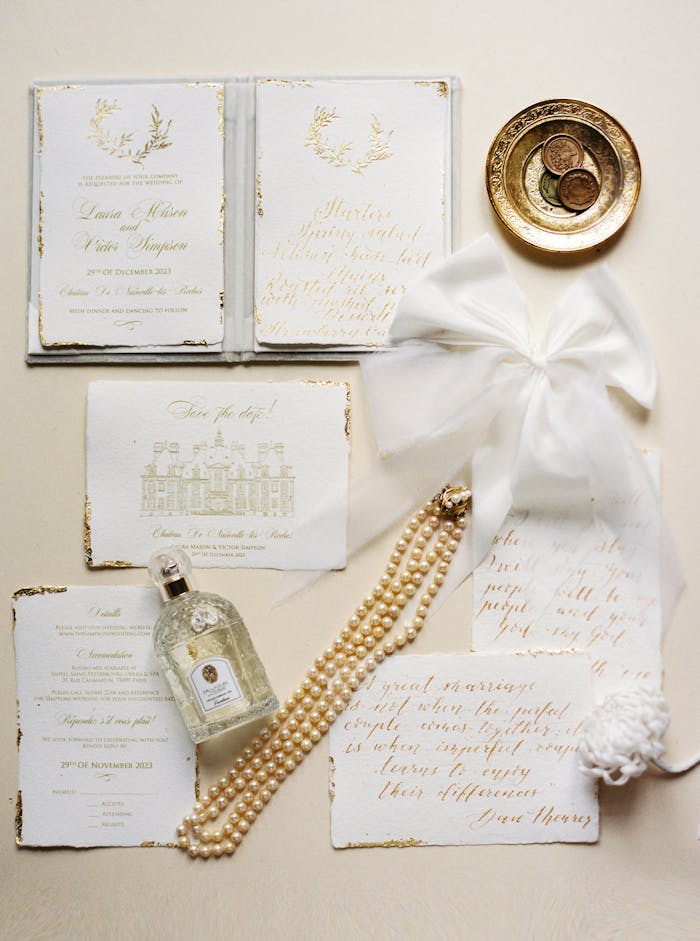 Sophisticated wedding stationery flat lay with perfume, pearls, coins, and a white ribbon on a neutral background.